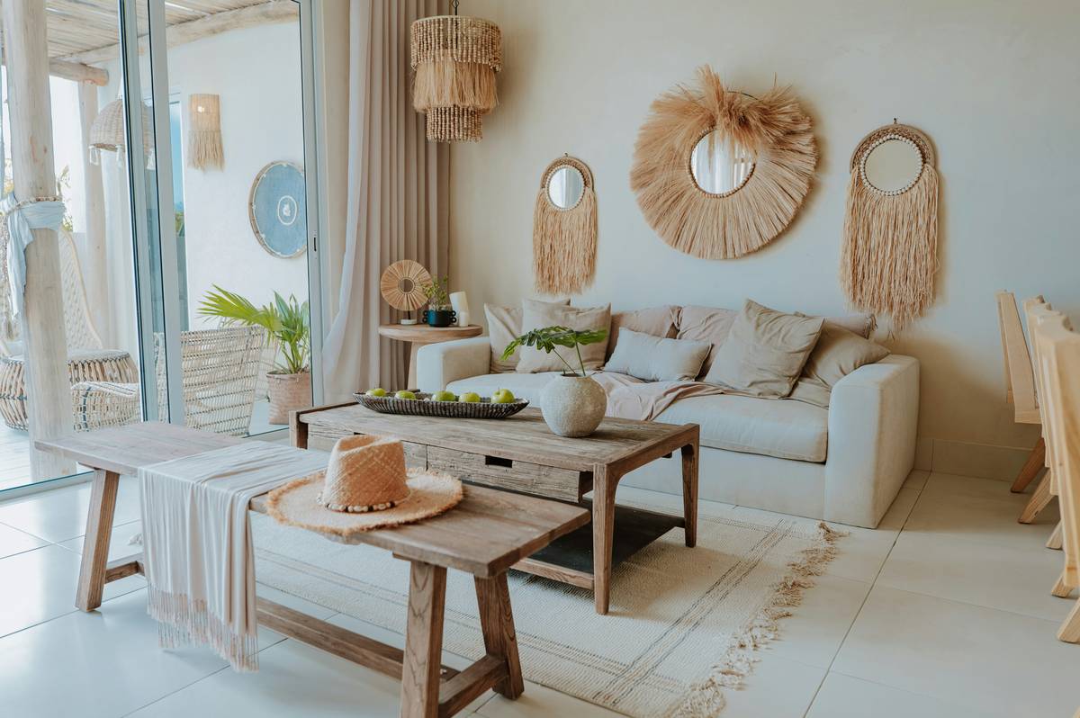 tropical style living room furniture including a light teak sofa, woven seagrass ottoman, and potted monstera in a bright, airy indoor space